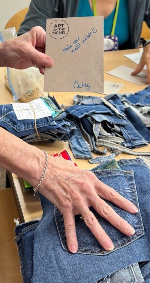 A hand holds a card that says 'ART ON THE MEND' and 'make your mend visible' next to stacks of denim fabric on a table.