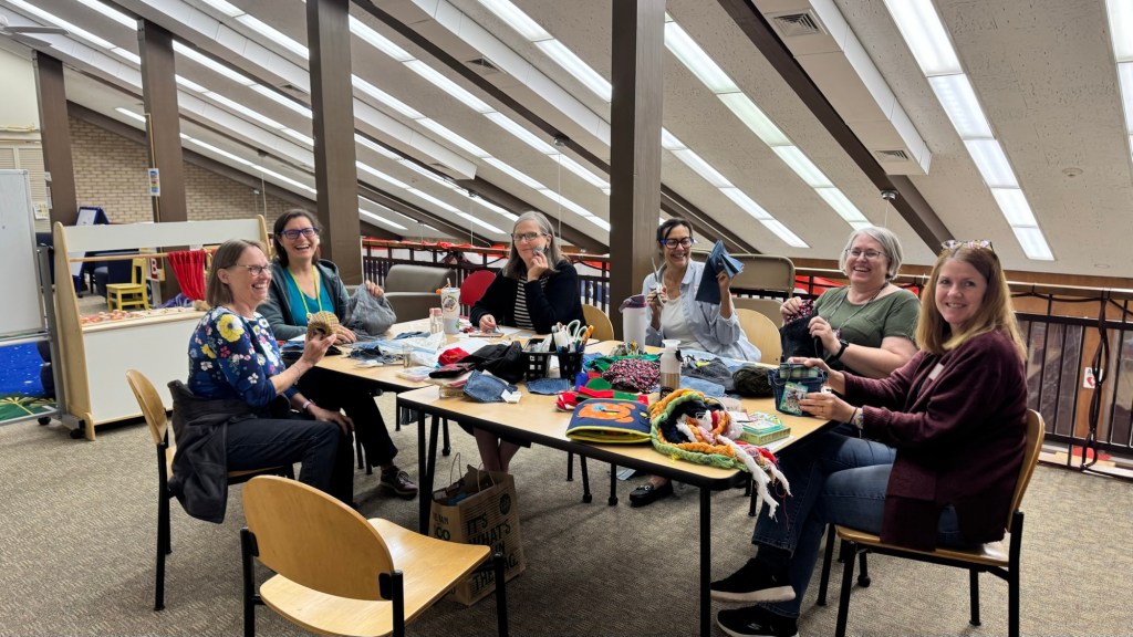 A group of six women sitting around a table in a brightly lit room, engaging in a craft activity with various materials and tools spread out on the table.
